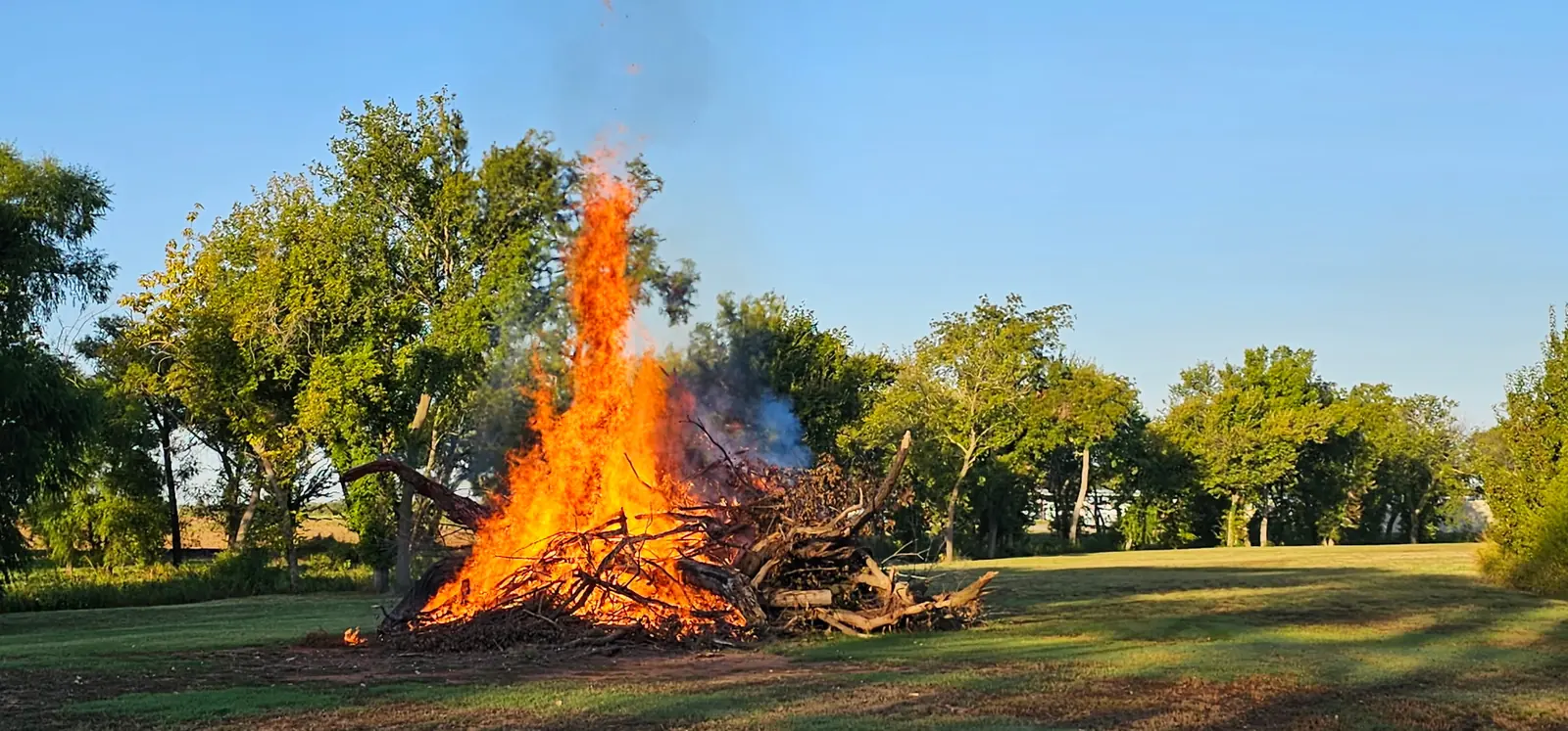 Brush pile burning with tall flames on a grassy lot beside a line of trees in Oklahoma