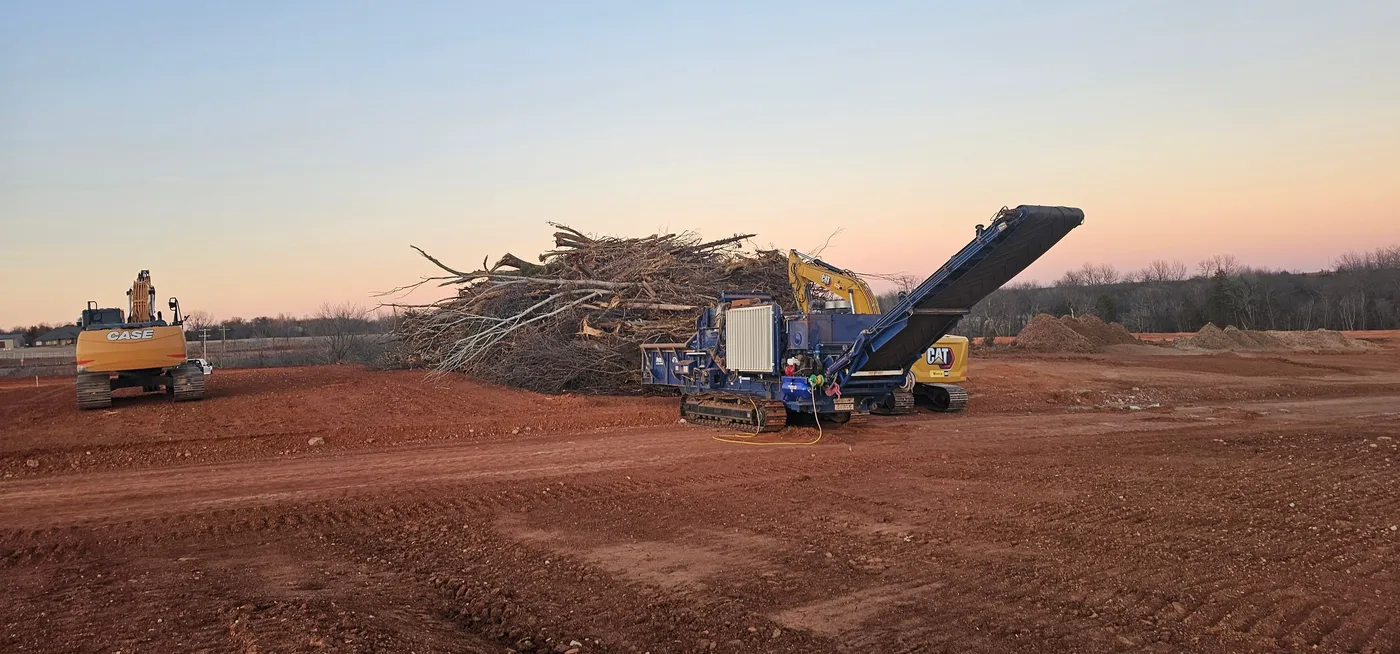 Grinder and excavators processing a large brush pile into mulch at dusk on an Oklahoma land clearing job