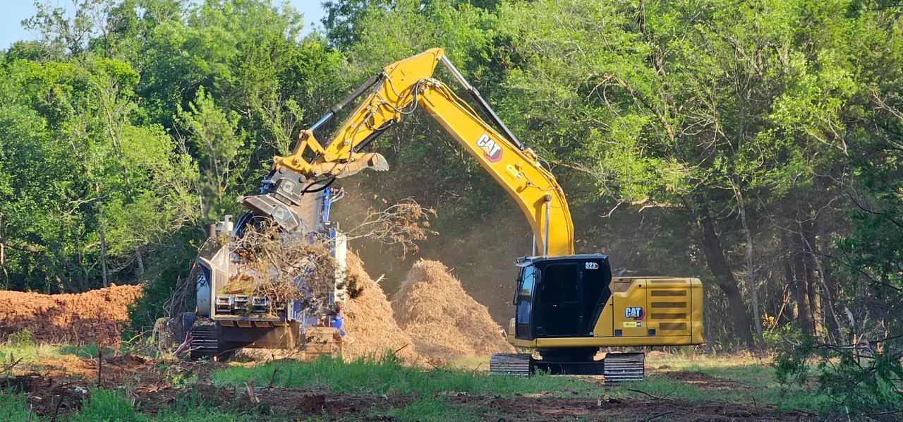 Cat excavator with a grapple feeding cleared brush into a grinder along an Oklahoma tree line