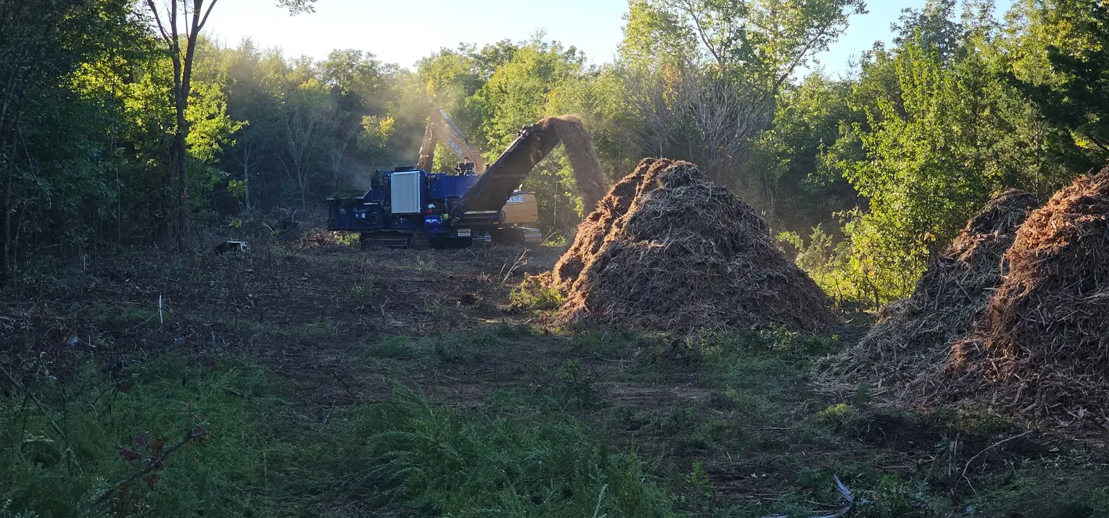 Horizontal grinder and excavator clearing a wooded lot beside a pile of fresh wood mulch on Oklahoma land