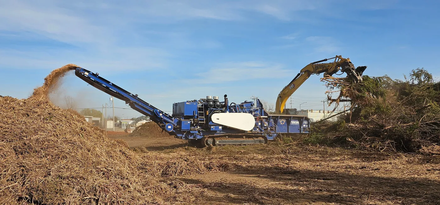 Horizontal grinder blowing wood mulch onto a pile as an excavator feeds it cleared brush near Oklahoma City